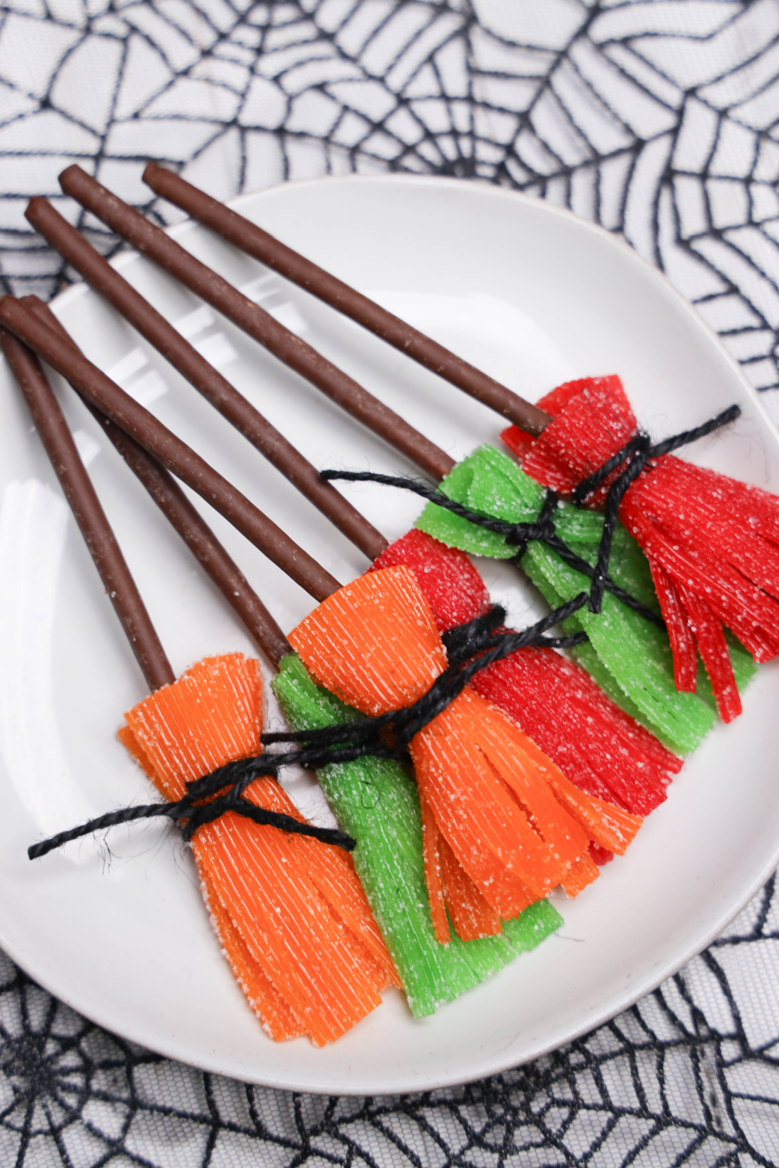 Orange, green, and red sour candy brooms with chocolate covered Pocky handles and black twine on a white plate.