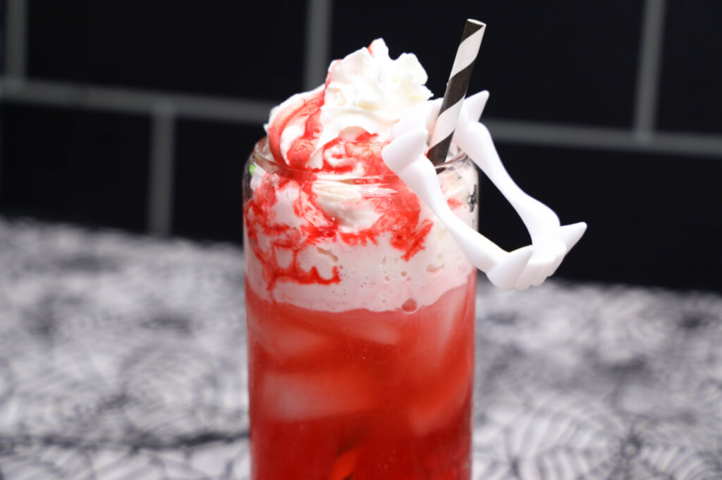 Red Vampire Dirty Soda over ice with whipped topping, strawberry syrup, a striped straw, and white plastic fangs.