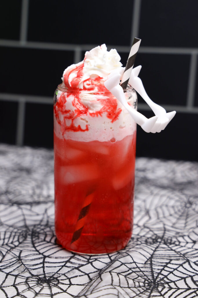Red Vampire Dirty Soda over ice with whipped topping, strawberry syrup, a striped straw, and white plastic fangs.
