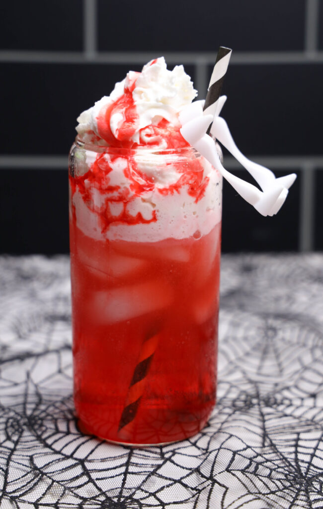 Red Vampire Dirty Soda over ice with whipped topping, strawberry syrup, a striped straw, and white plastic fangs.